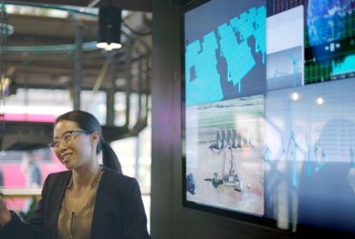 Stock photograph of a young Asian woman conducting a seminar / lecture with the aid of a large screen. The screen is displaying data & designs concerning low carbon electricity production with solar panels & wind turbines. These are juxtaposed with an image of conventional fossil fuel oil production.